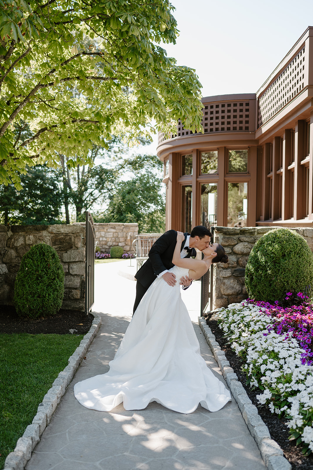 Groom dipping and kissing the bride on a stone garden path lined with vibrant flowers.